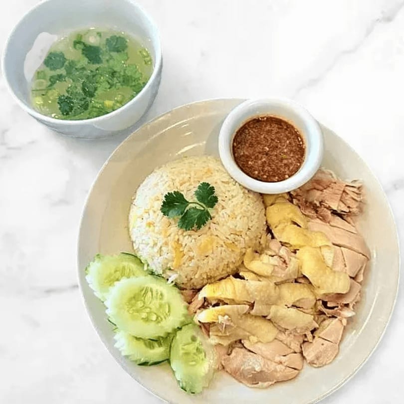 Thai chicken rice with sliced cucumbers, chili sauce, and a side of clear soup.