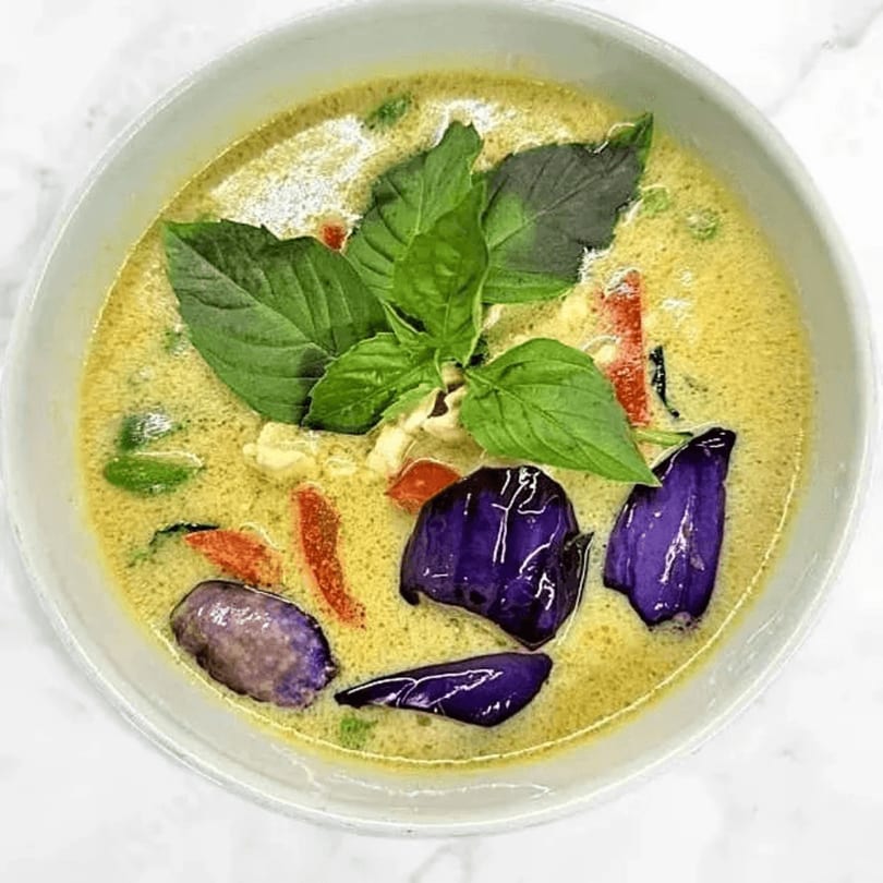Red Jasmine Thai green curry with purple eggplant, red peppers, and fresh basil in a white bowl.