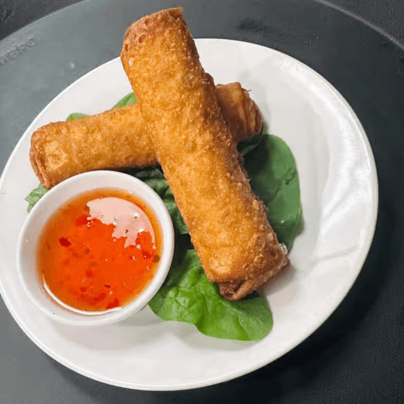 Two crispy Thai spring rolls on a bed of spinach with a side of sweet chili dipping sauce.
