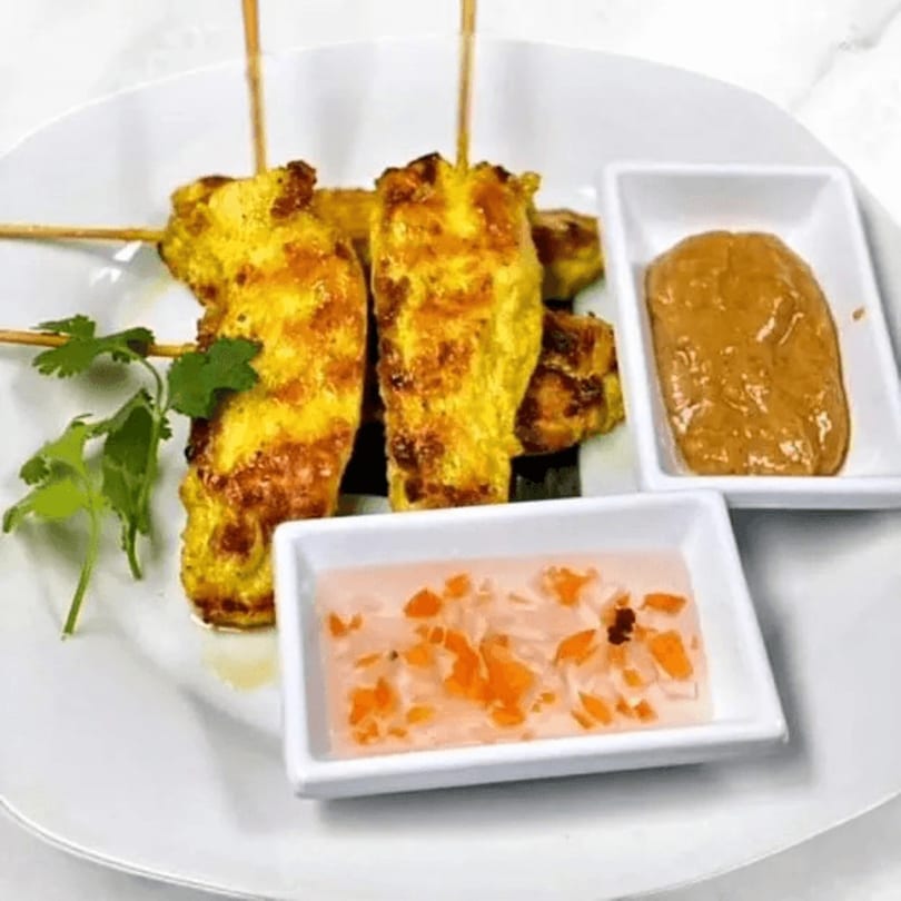 Chicken satay skewers with peanut sauce and sweet chili sauce on a white plate.